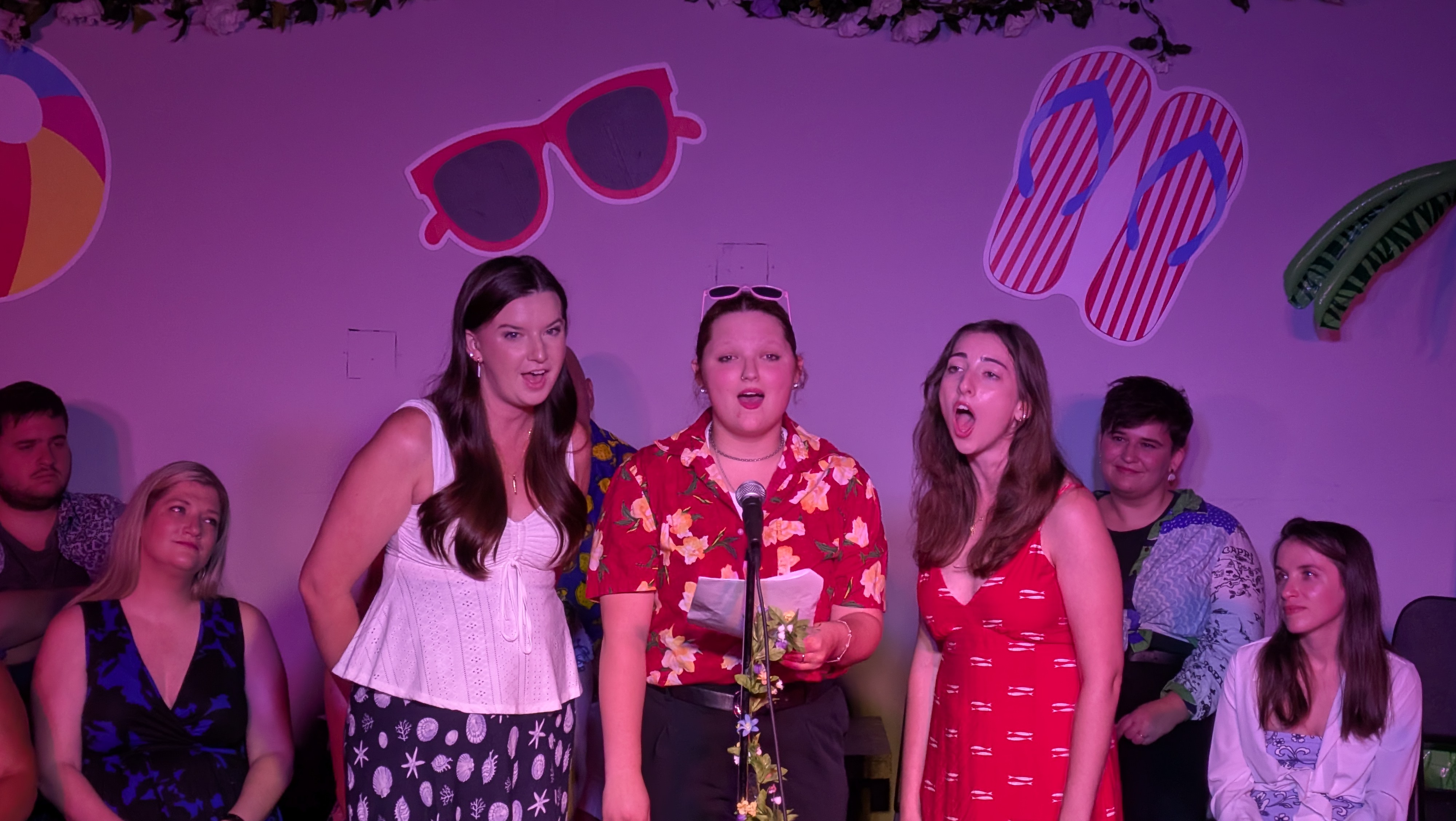 Three singers around one microphone. The stage is lit pink, with a white light illuminating the singers.