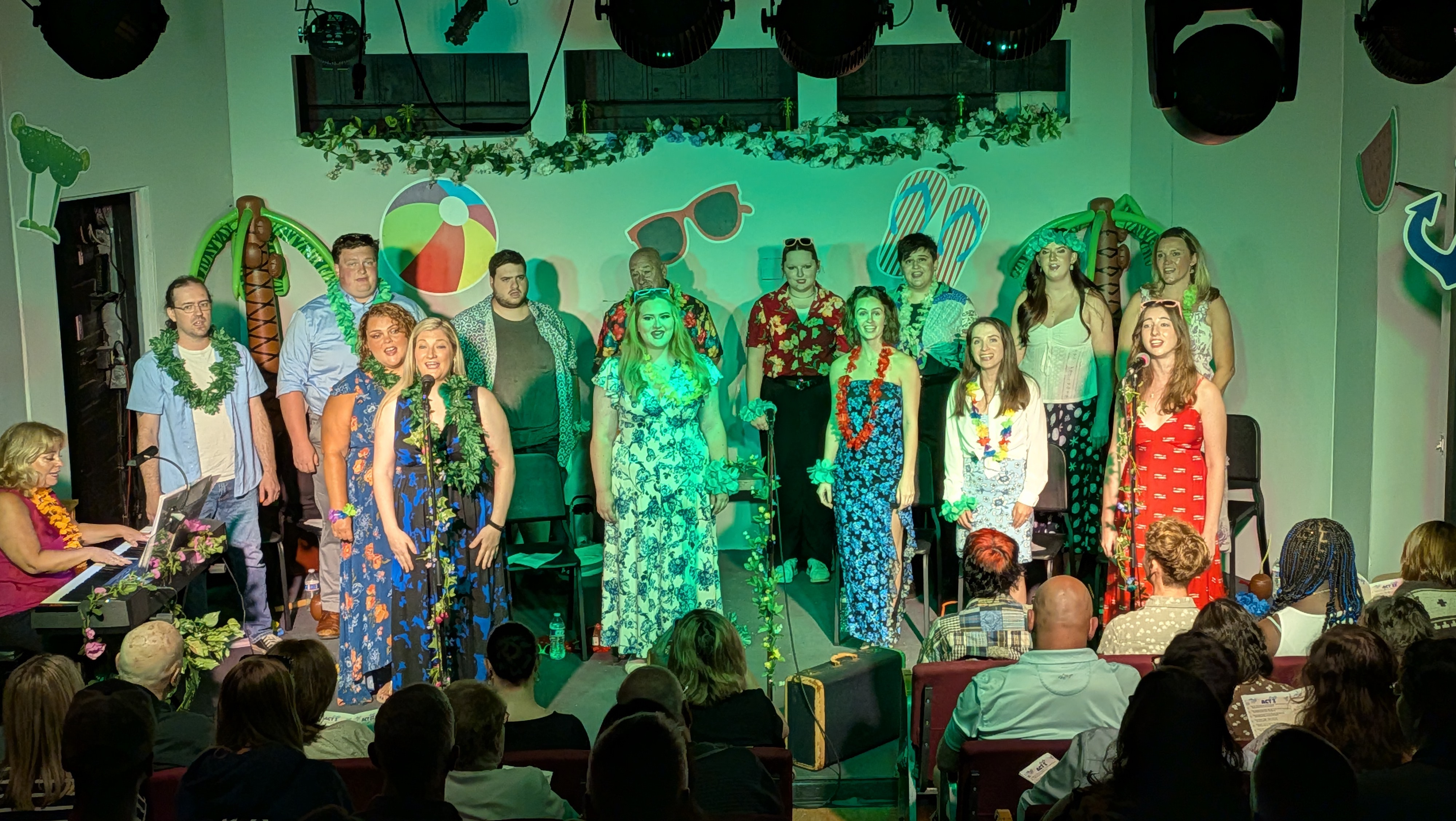 The entire cast of the cabaret standing. One singer is at the SL microphone, while another singer is at the SR microphone. The stage is lit in green lights, with the two soloists being illuminated by white lights.